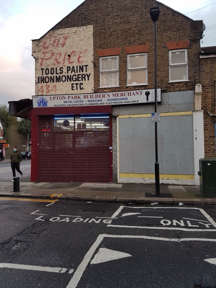 The gable end above Upton Park Builders Merchants showing the faded hand-painted heritage sign reading Cut Price Tools, Paint, Ironmongery, above the shop's own sign for metal gates, windows and signboards.