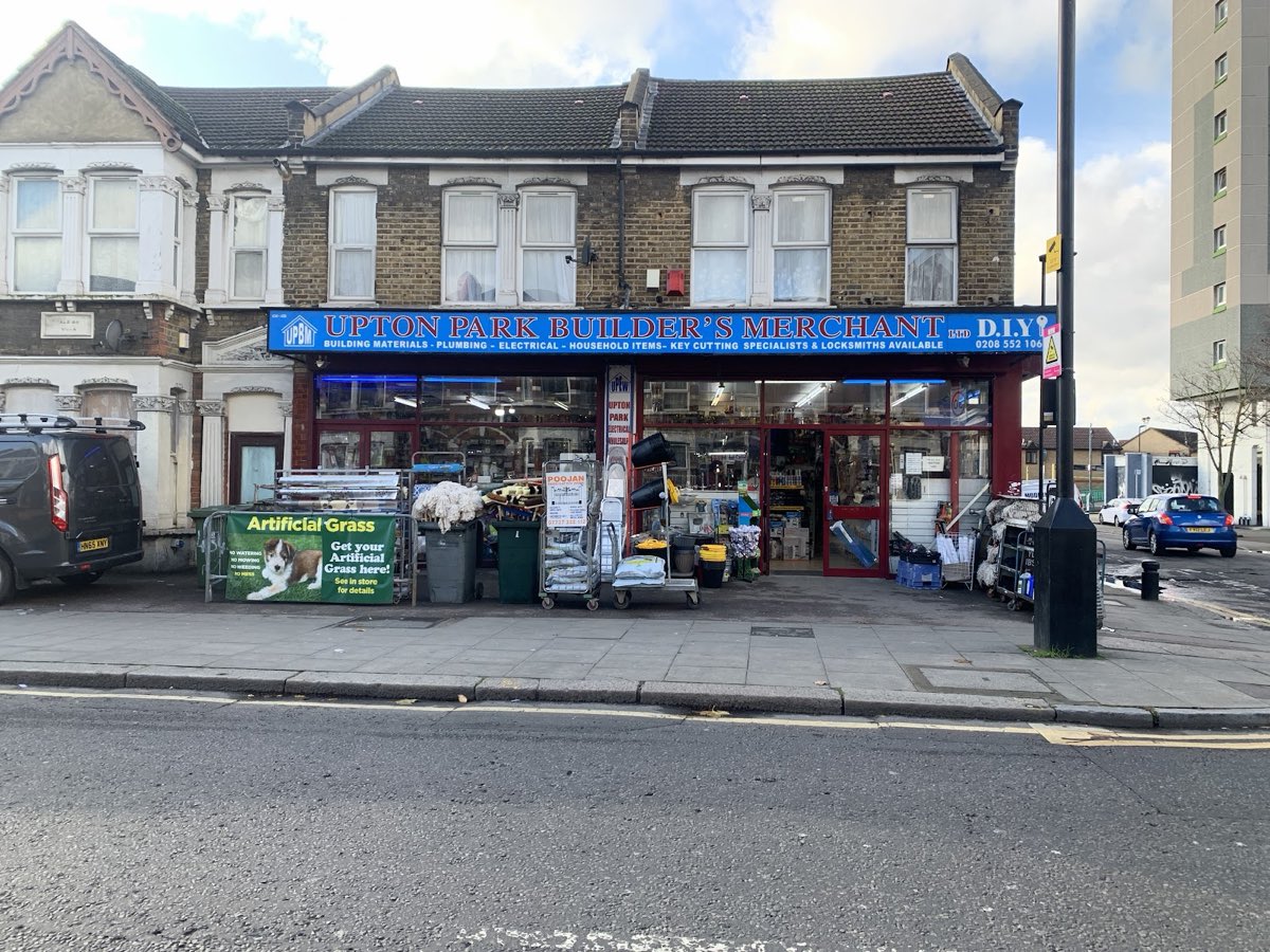 The blue and red shopfront of Upton Park Builders Merchants at 434-436 Green Street, London E13, its fascia reading Building Materials, Plumbing, Electrical, Household Items, Key Cutting Specialists and Locksmiths Available.